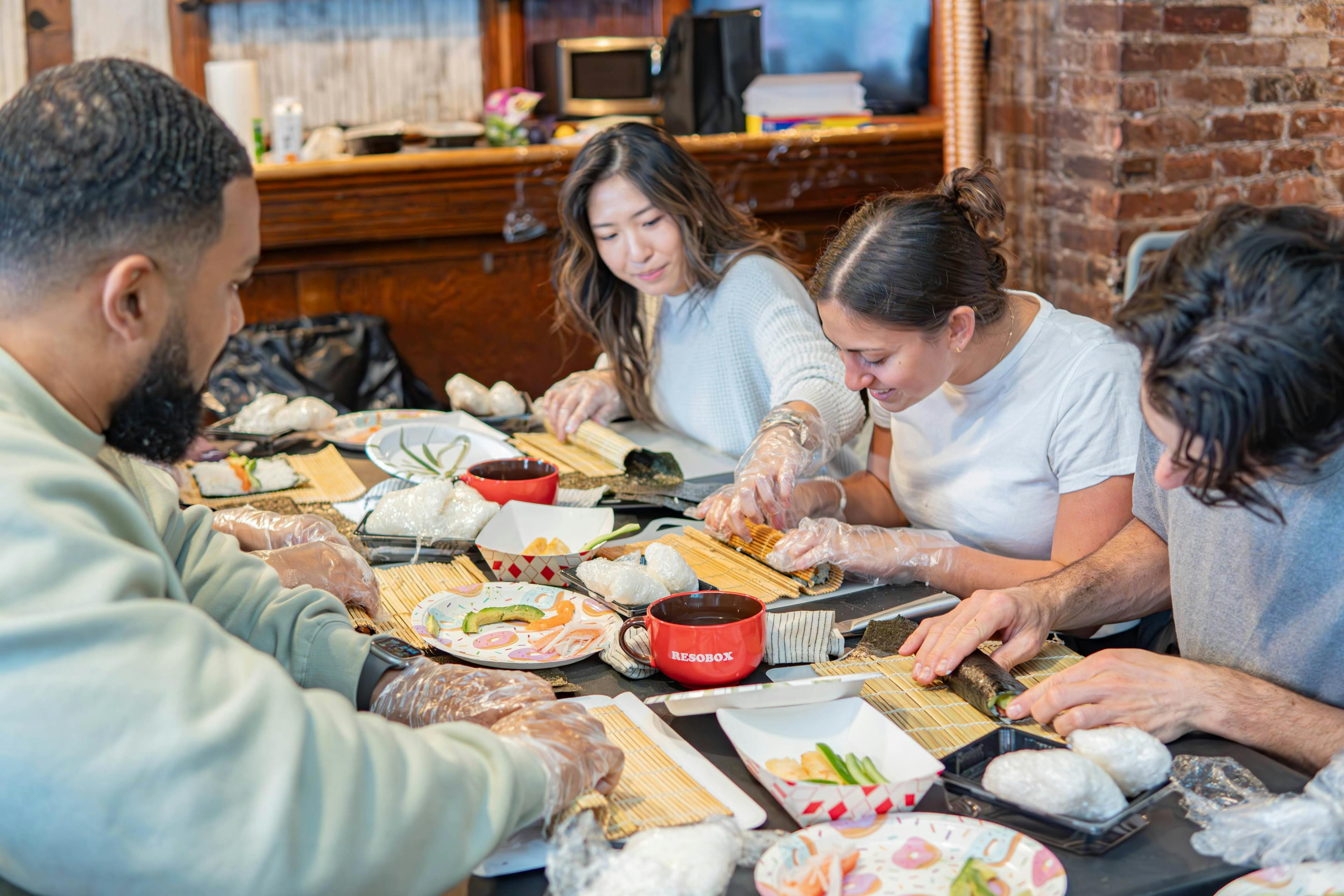 Four adults making sushi together using bamboo mats and various ingredients on a wooden table.