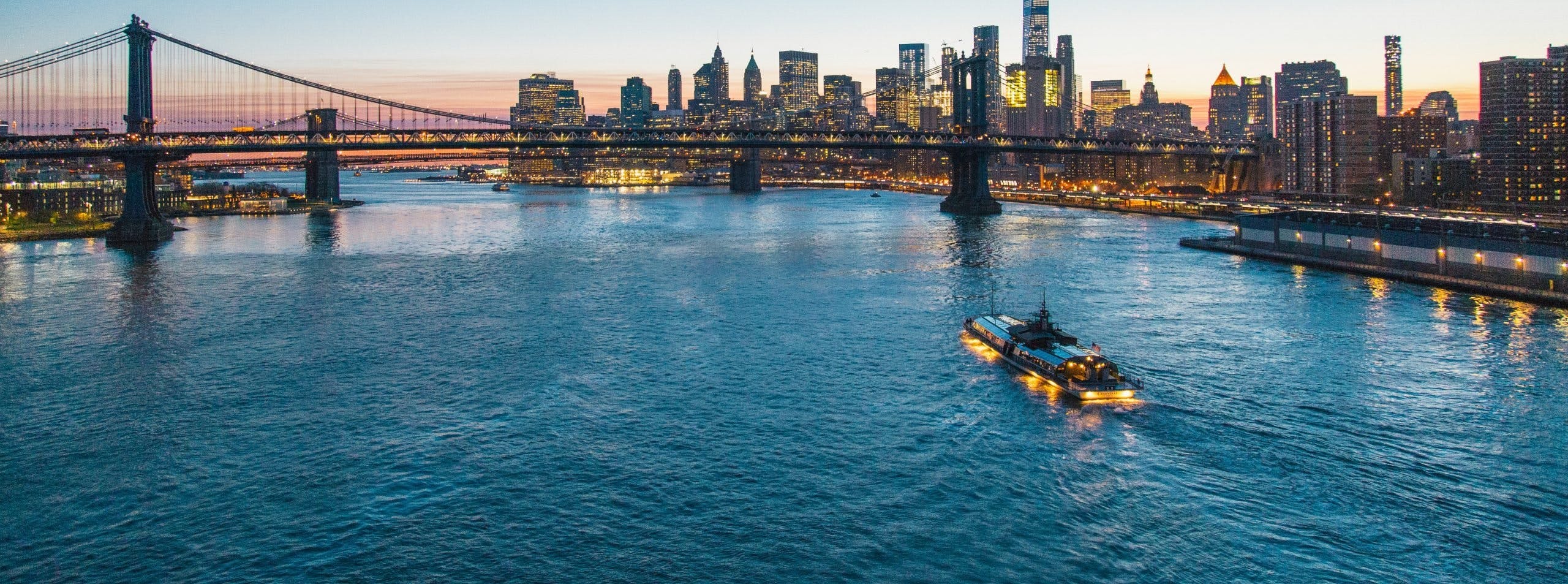 This image showcases a serene evening scene with a private boat gracefully cruising on the water, with the majestic New York City skyline and the illuminated Brooklyn Bridge in the background. It captures the essence of an exclusive Manhattan boat cruise experience, offering a tranquil escape from the bustling city life while presenting an enchanting view of the urban landscape as day turns to night.