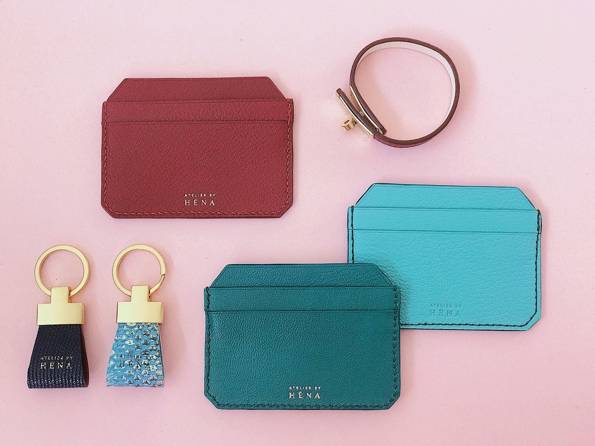 Assorted leather goods on pink: maroon card holder, two keychains, dark turquoise wallet, teal card holder, and a thin brown belt.
