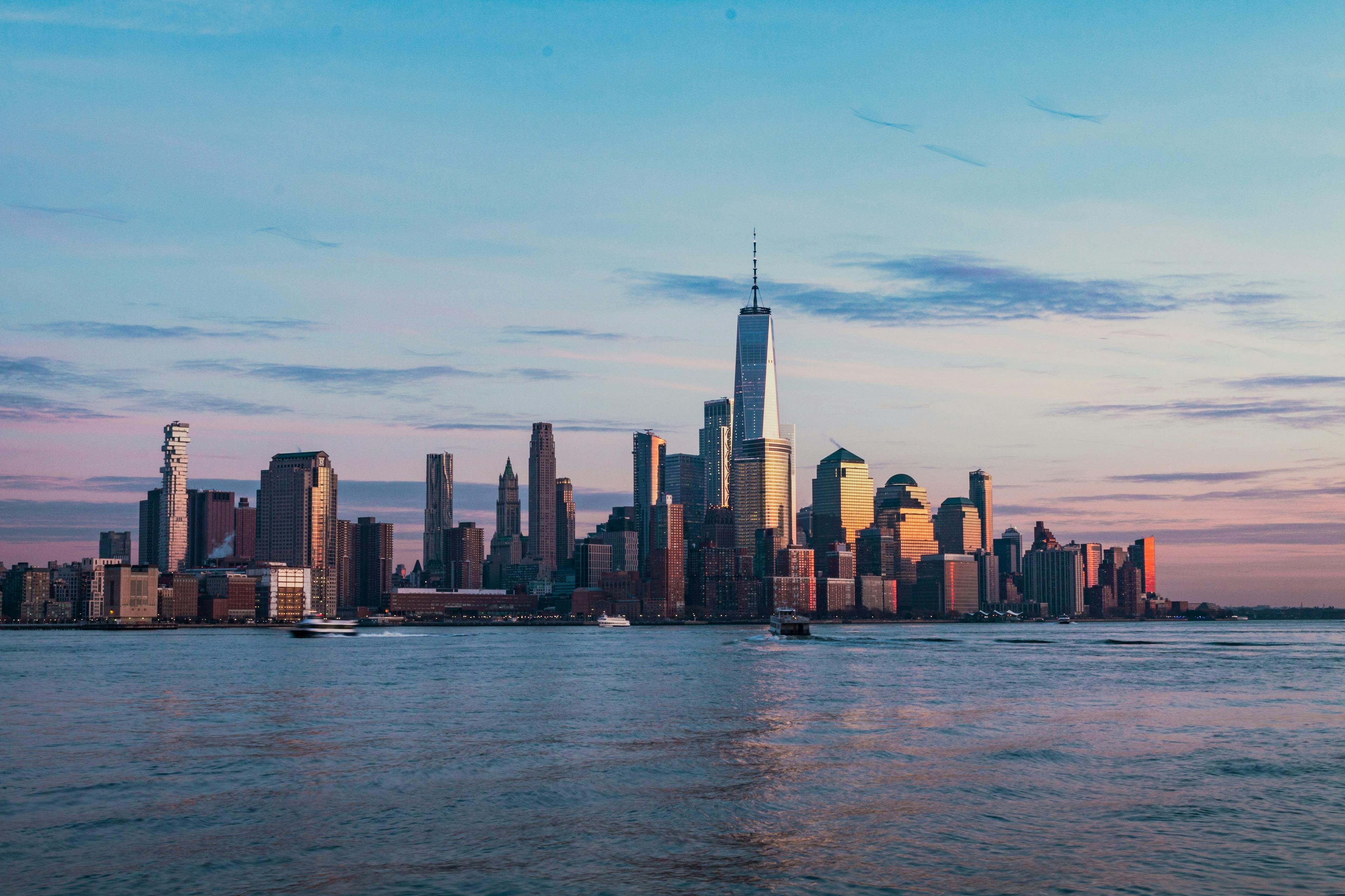 This image captures a serene view of the New York City skyline during sunset. The sky is painted with soft pink and blue hues, providing a picturesque backdrop to the iconic skyscrapers, including the One World Trade Center. The river in the foreground reflects some of the colors of the sky, enhancing the overall tranquility of the scene. Small boats can be seen on the water, adding a dynamic element to the peaceful landscape. This image highlights the beauty of the city's architecture contrasted with the natural elements of the sky and water during the golden hour.