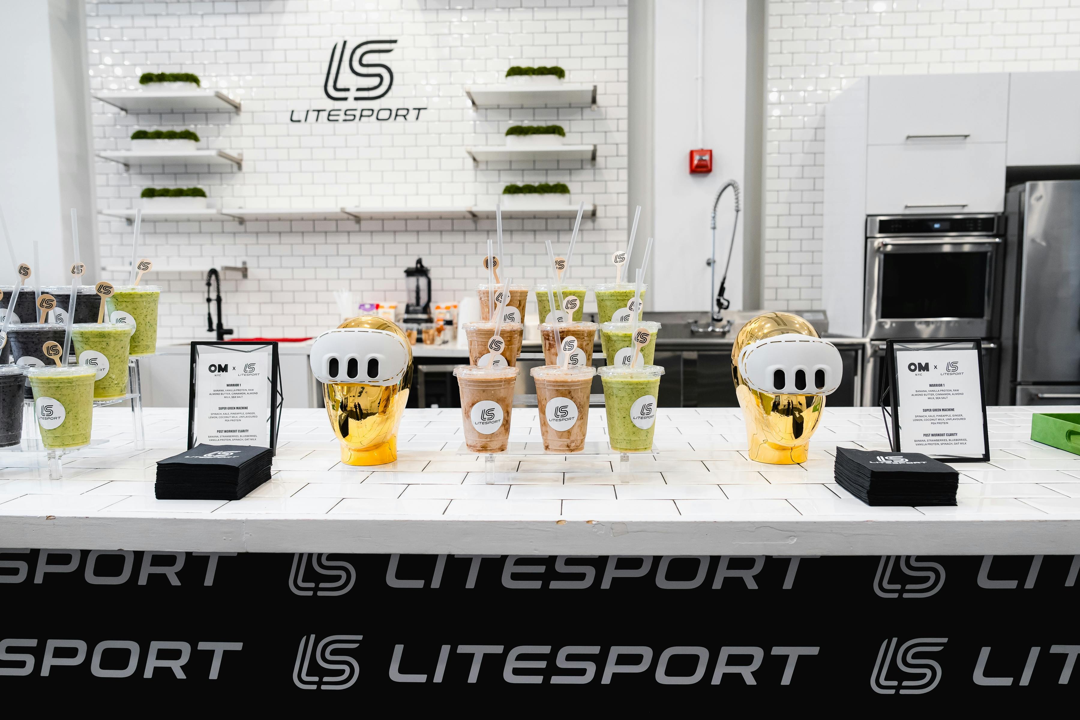 Interior view of the LiteSport smoothie bar featuring a variety of green and brown smoothies in clear cups with lids and straws, displayed on a white tiled counter. Prominently placed are two gold trophies and a black sign reading 'OM x LS. LiteSport x Omni Smiles'. The backdrop is a white, clean kitchen space with a logo of LiteSport on the wall.