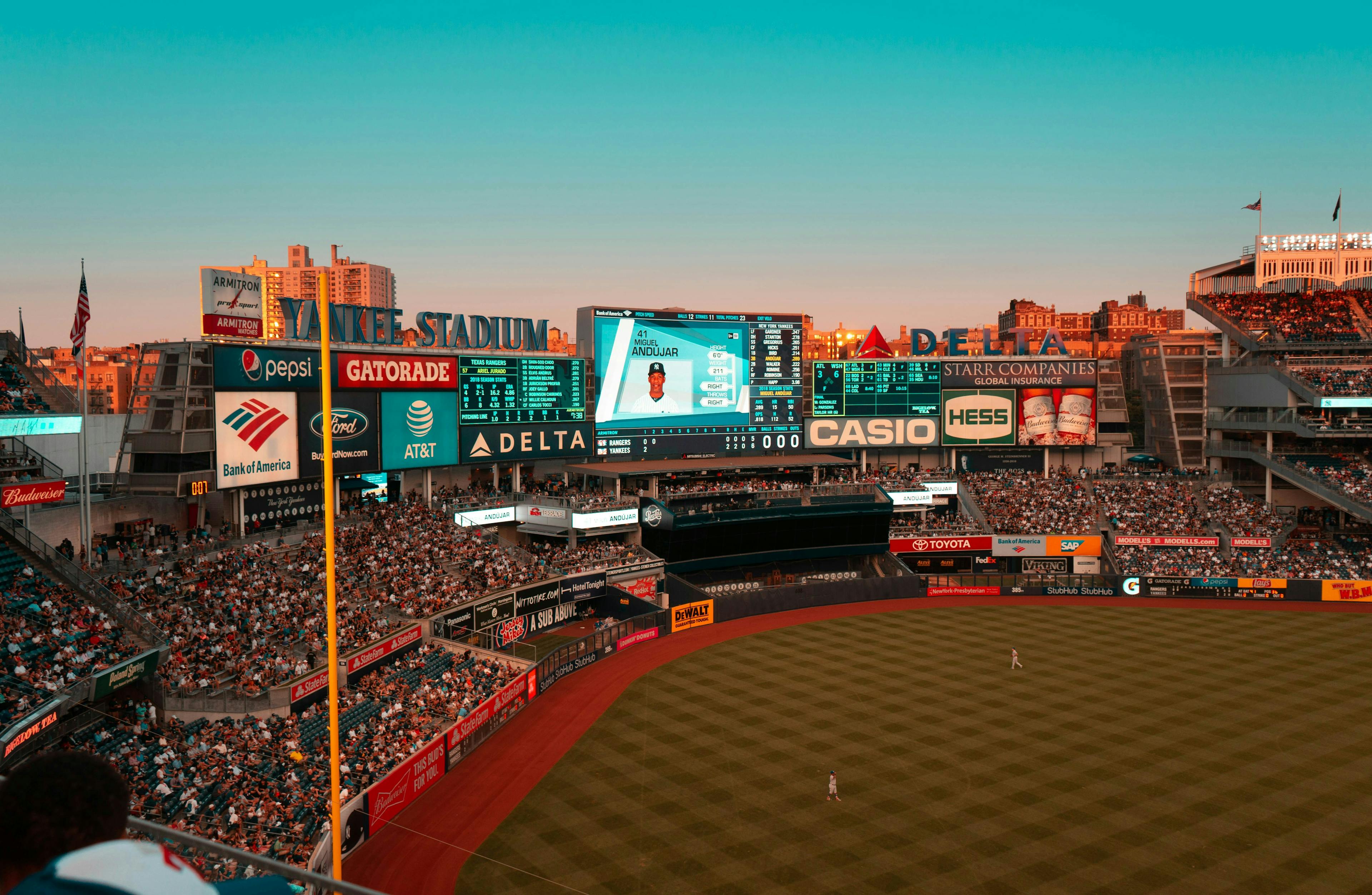 View of a baseball game at Yankee Stadium during a warm sunset, showing the field, crowded stands, and the large digital scoreboard displaying player information and advertisements.