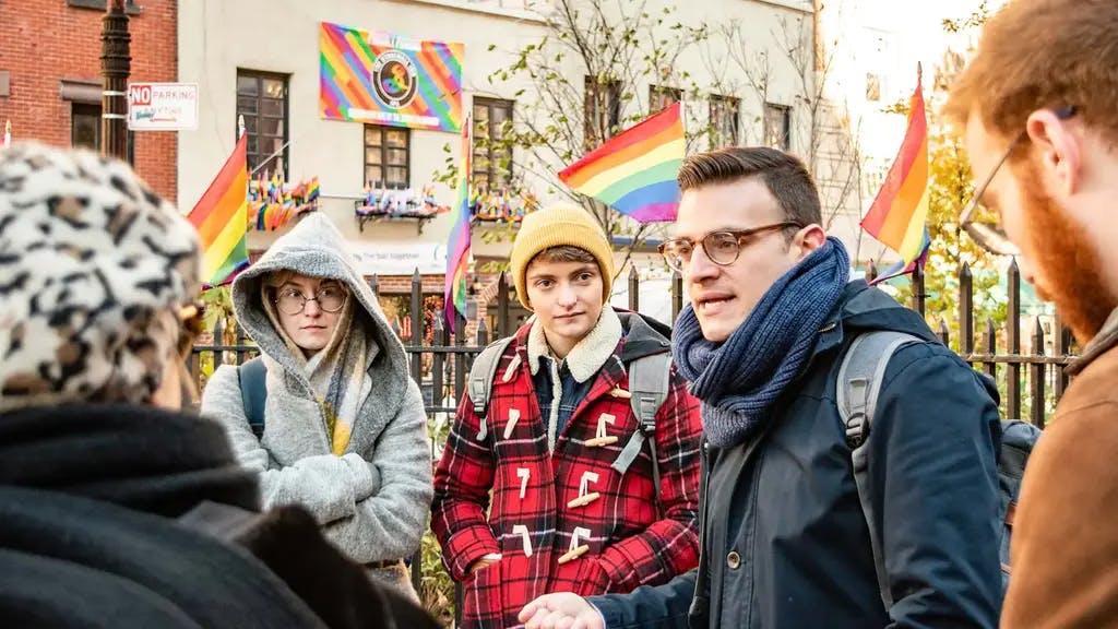 a man talking to a group of people with pride flags on buildings in the background