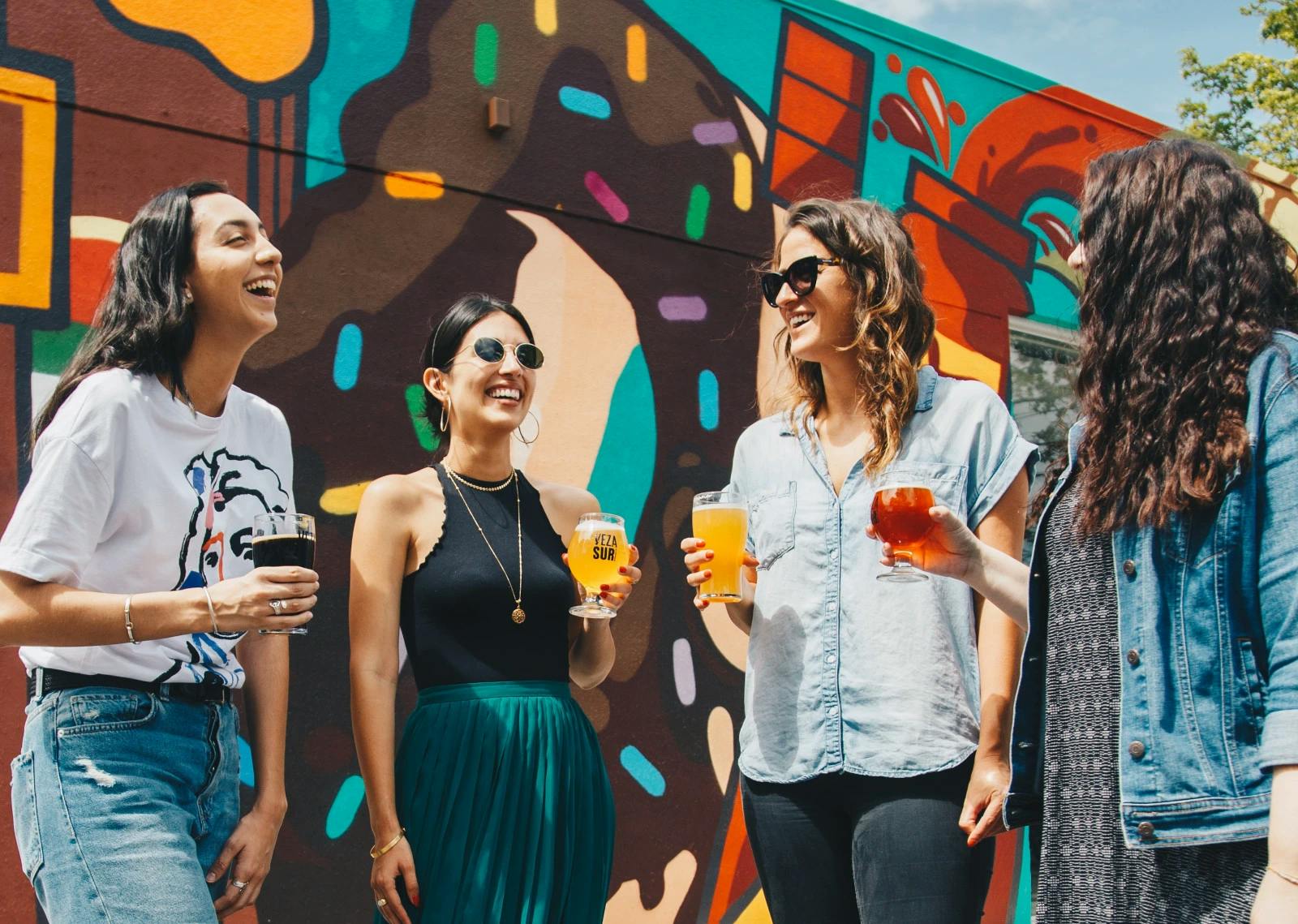 a group of women enjoying a drink and laughing in front of a colorful graphity wall