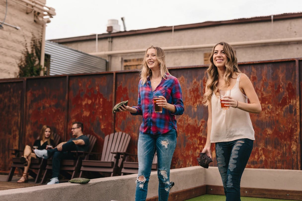 two women laughing while playing corn hole and having a drink
