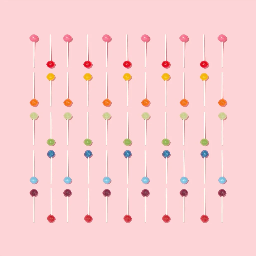 Neatly arranged rows of colorful lollipops on a pink background, each with a different vibrant hue, creating a cheerful and playful display.