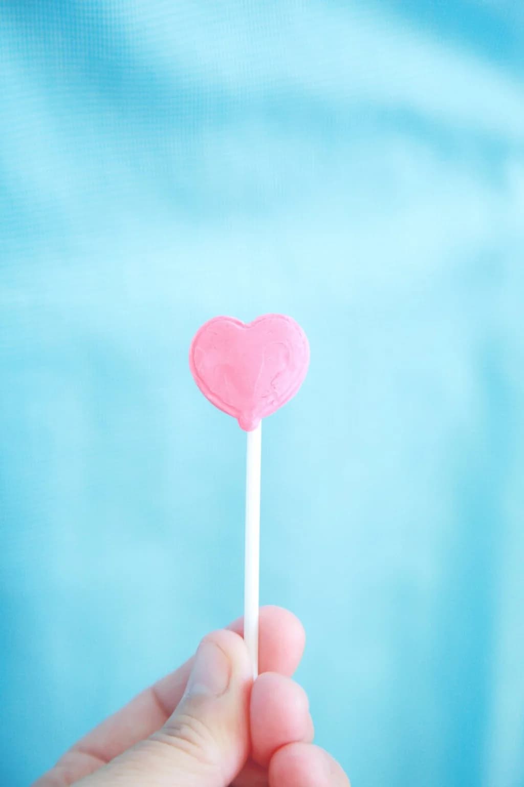 A close-up of a pink heart-shaped lollipop being held against a soft, pastel blue background, evoking a sense of sweetness and love.