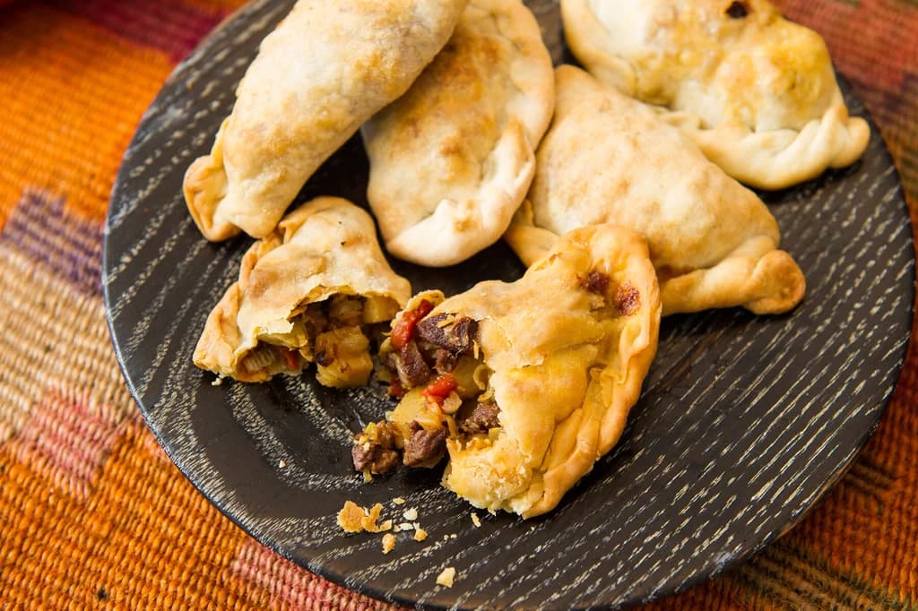 A plate of golden-brown empanadas with one broken open to reveal a savory filling of meat, vegetables, and spices, set on a colorful woven cloth.