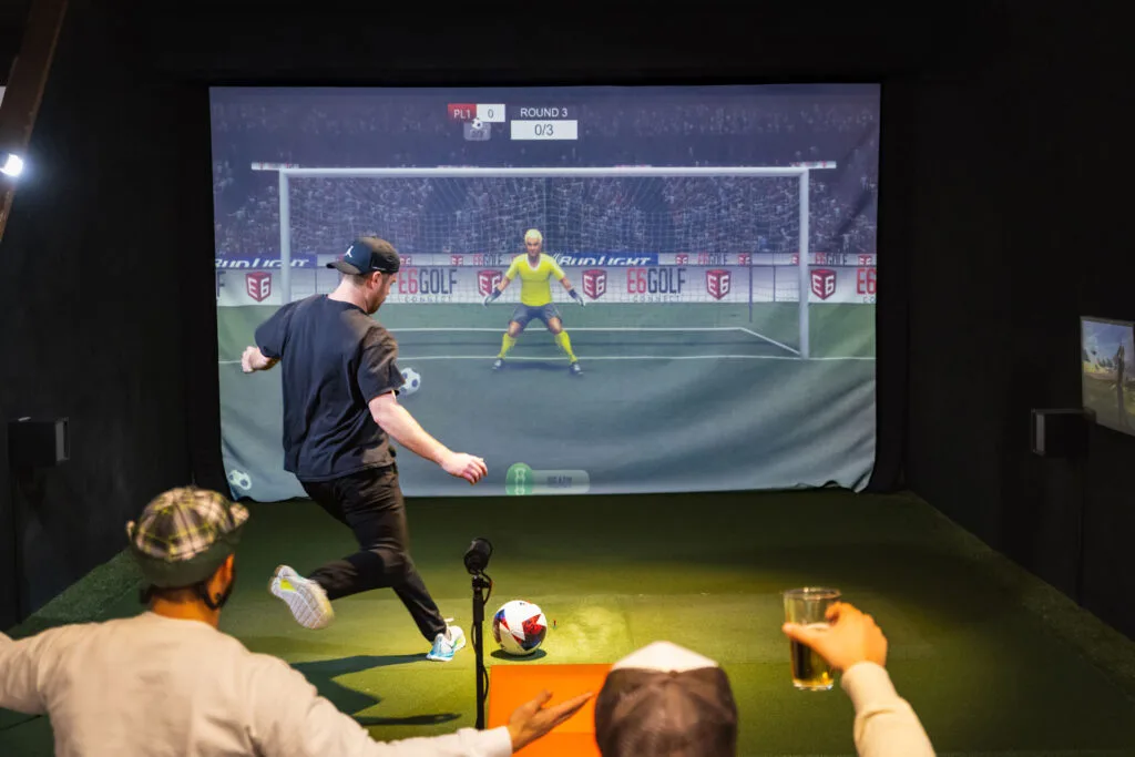 A group of people enjoying a soccer simulation game, with one person kicking a ball towards a digital goal on a large screen.