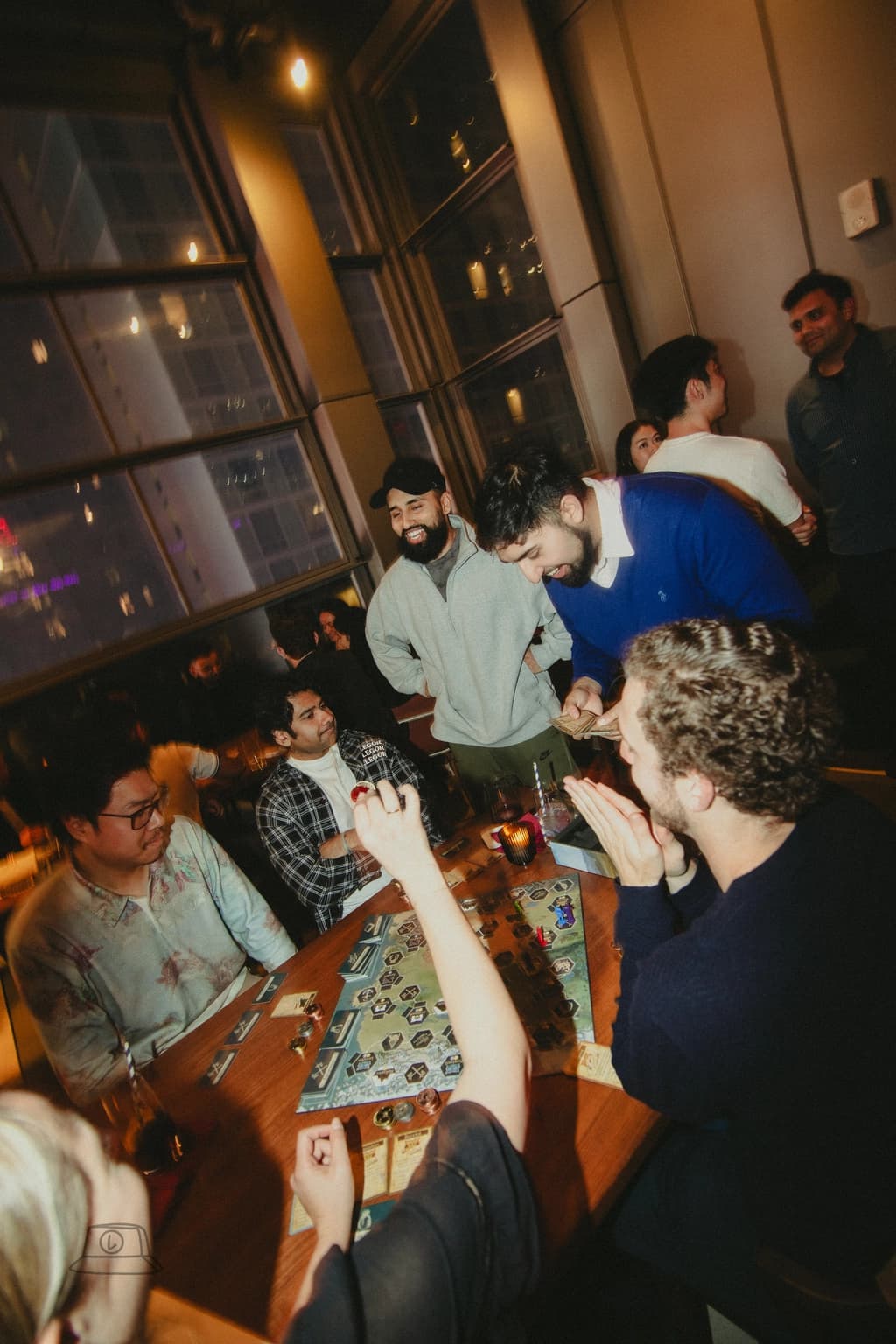 A group of friends intensely focused on a board game while others observe and enjoy drinks.