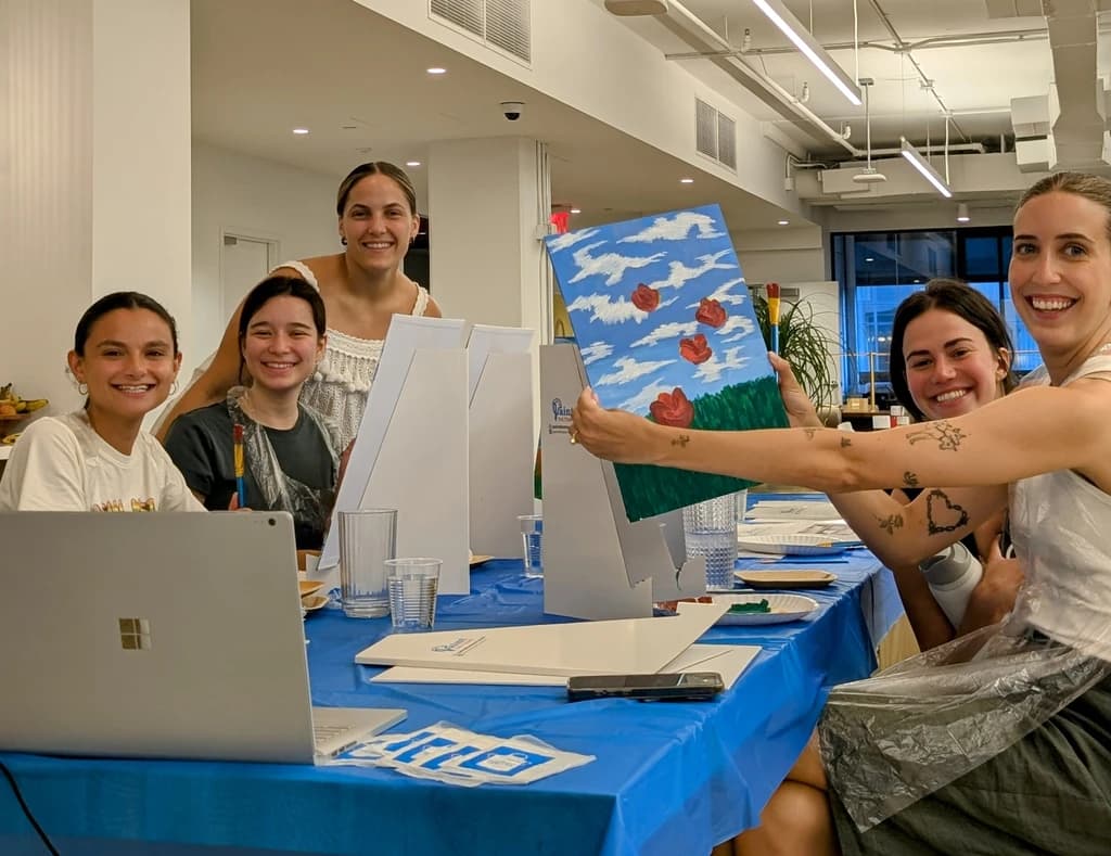 A group of five smiling women participating in a painting class, holding up their artwork with pride, surrounded by easels and paint supplies.