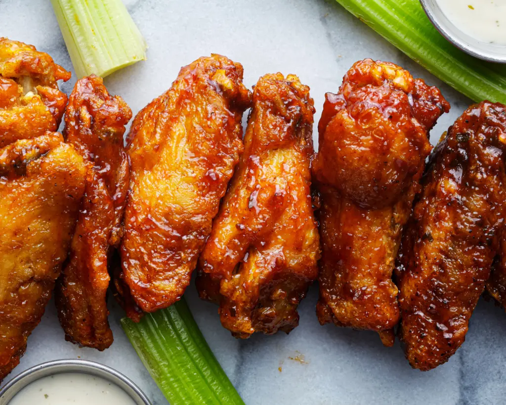 Close-up of golden-brown chicken wings glazed with spicy sauce, accompanied by celery sticks and dipping sauces on the side.
