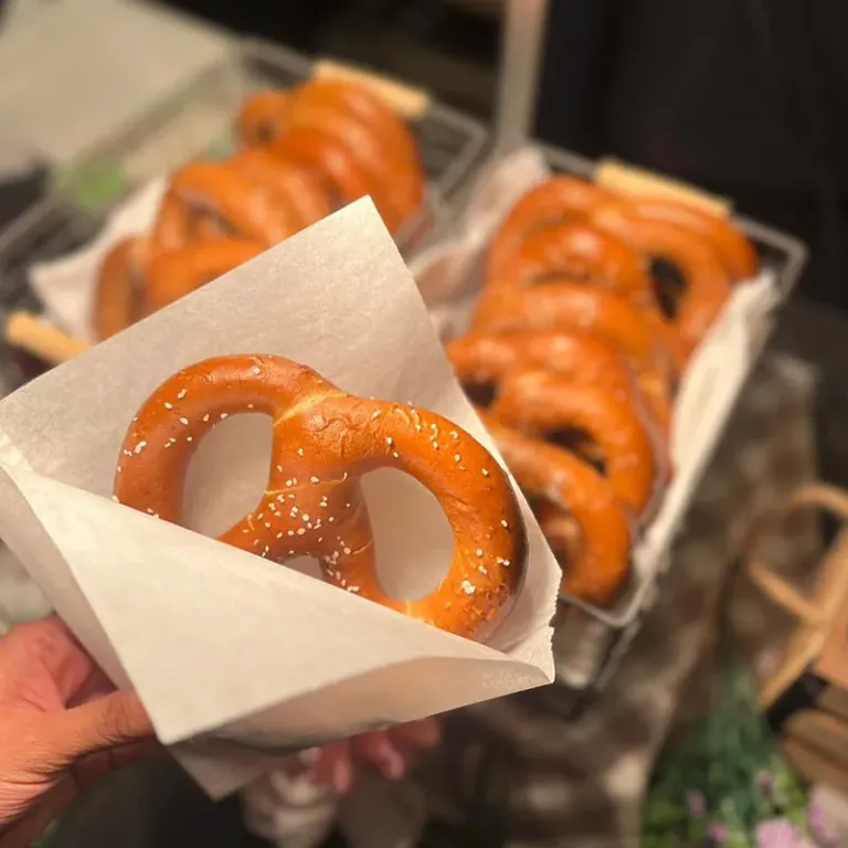 Hand holding a salted soft pretzel in a paper sleeve, with a tray of pretzels in the background.