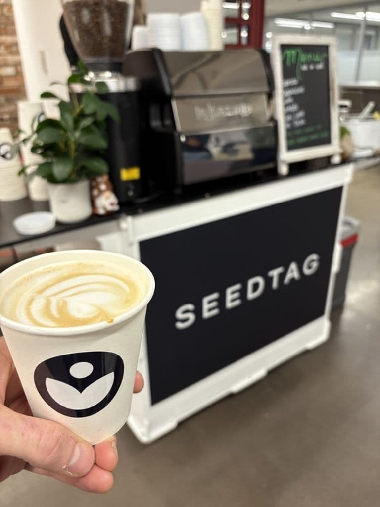 A hand holds a cup of latte with latte art in front of a mobile coffee cart branded "SEEDTAG" with an espresso machine and menu board.