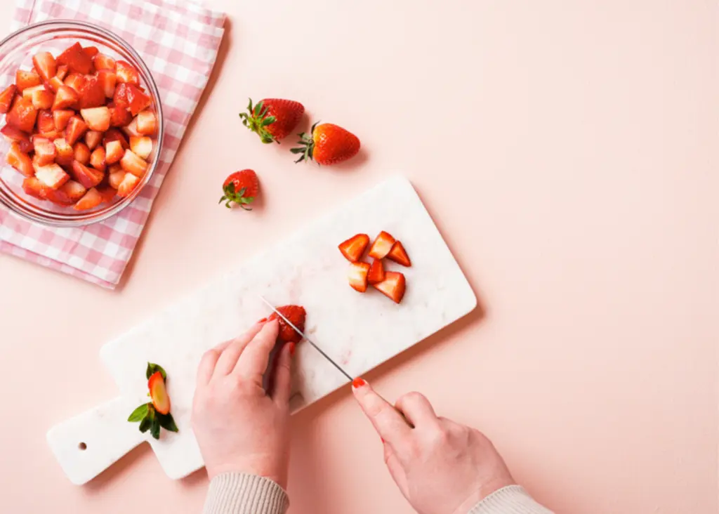 A close-up of hands chopping fresh strawberries on a marble cutting board. A bowl of diced strawberries sits nearby, along with a few whole strawberries scattered on a pink checkered cloth.
