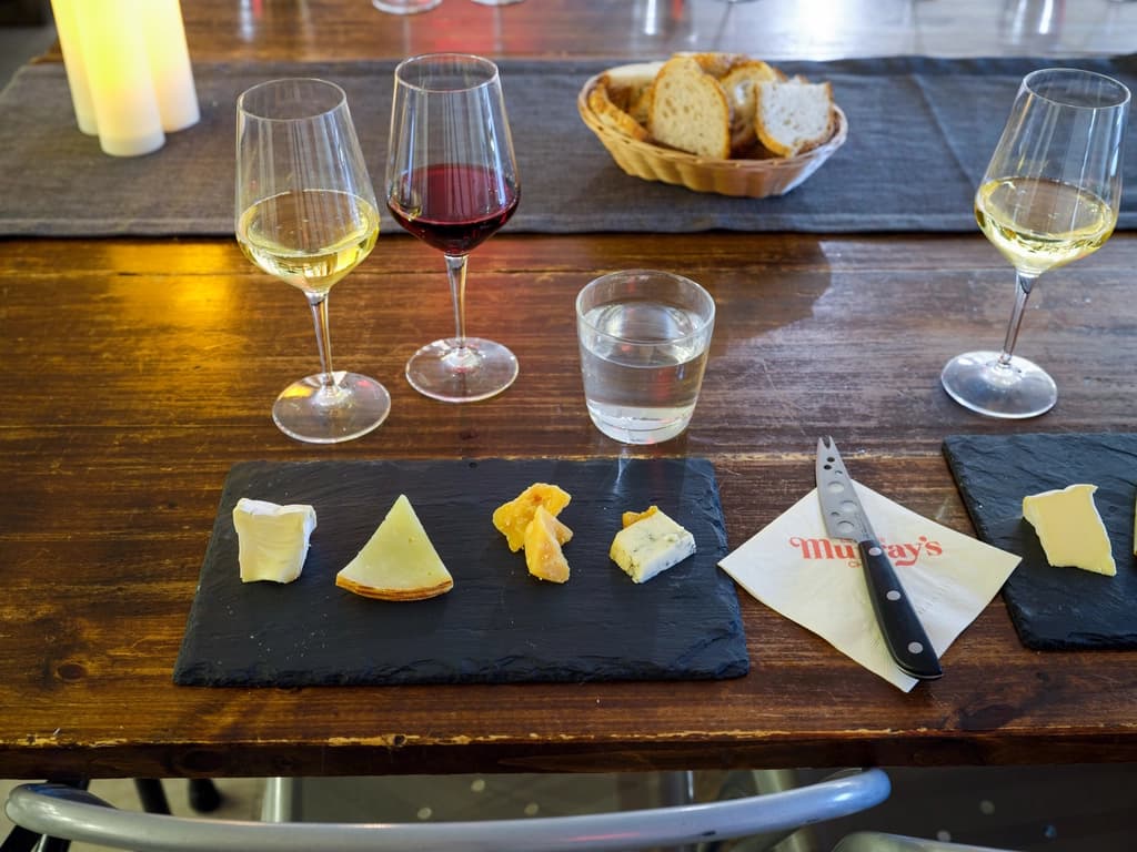 A cheese tasting setup with three glasses of wine, a slate with various cheese samples, and a basket of bread on a wooden table.