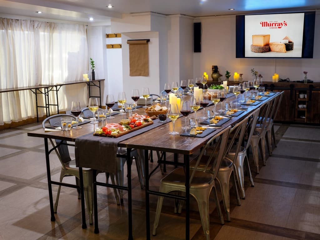 A long table set for a cheese tasting event, with glasses of wine, cheese samples, and a screen displaying 'Murray's Cheese' in the background.