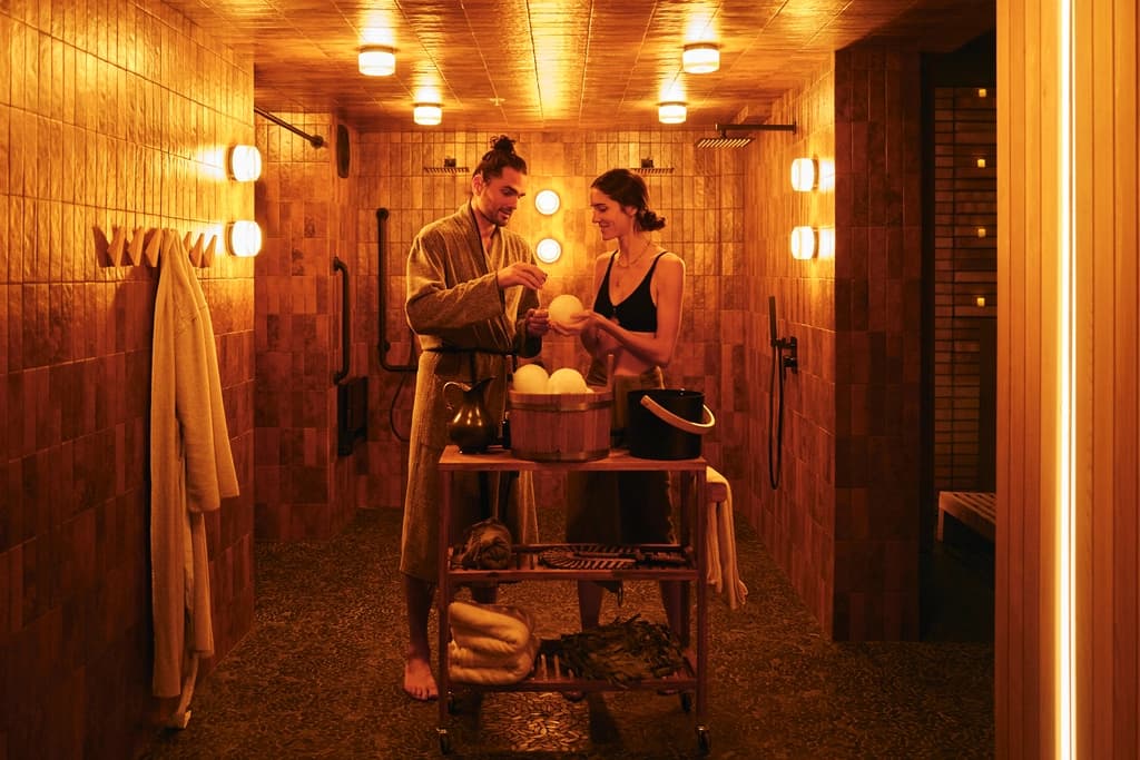 A man and woman preparing to enjoy a sauna ritual, surrounded by warm lighting and wooden spa tools, with towels and robes neatly arranged in a cozy, tiled room.