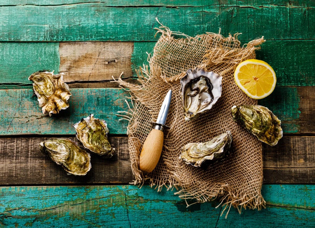 Hands opening an oyster with a knife, surrounded by lemon wedges and ice on a dark background.