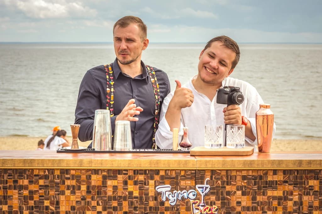 Two men behind a bar on a beach, one giving a thumbs-up while holding a camera