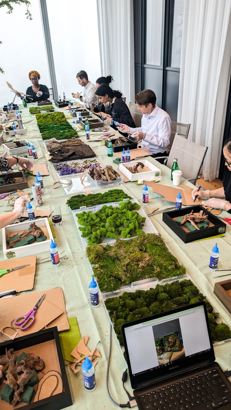 A group of people gathered around a long table in a modern space, creating moss wall art using glue, moss, bark, and wood pieces.