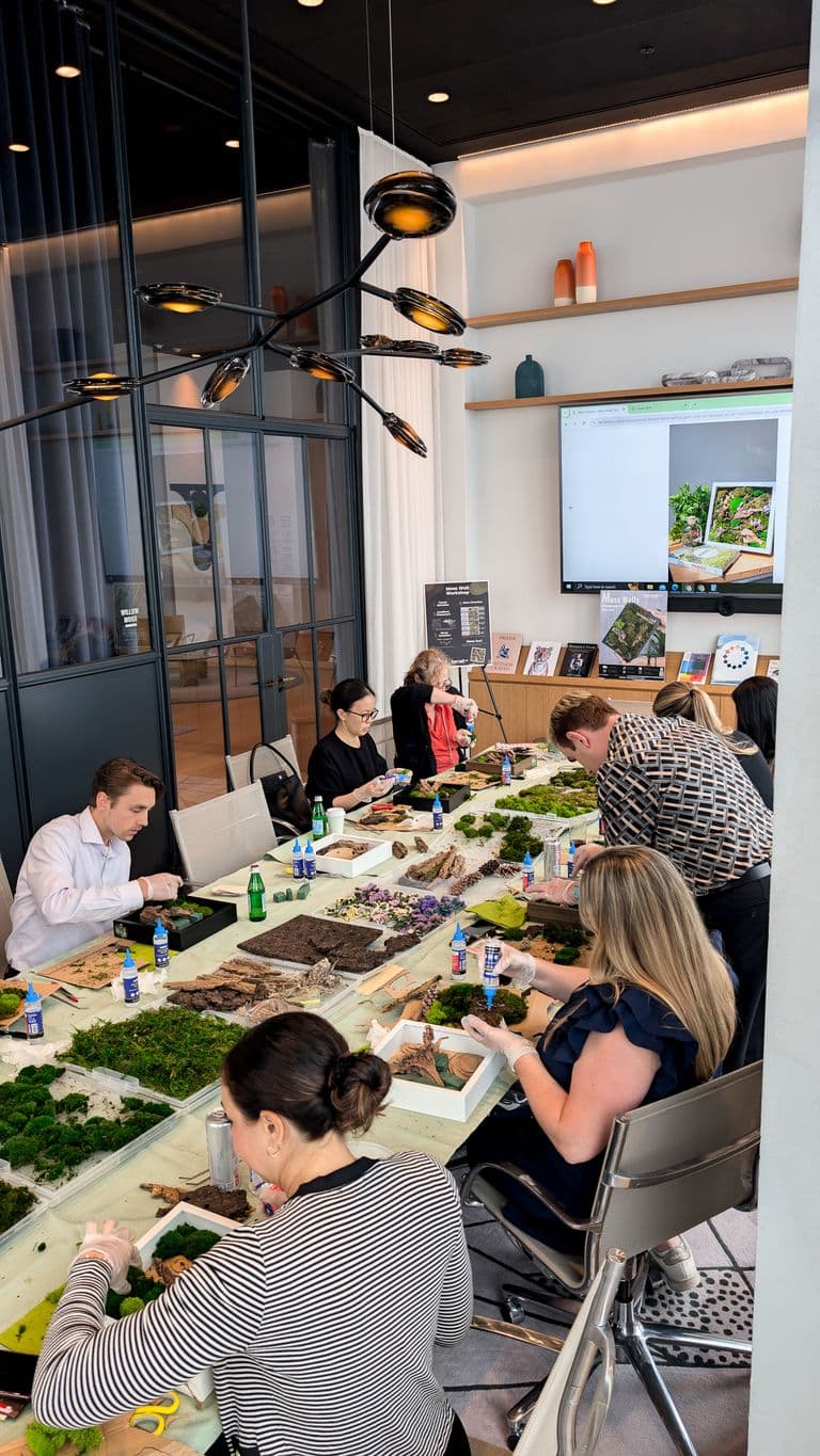 Participants at a team-building workshop assembling moss wall art, with trays of moss, glue bottles, and crafting tools on a long table.