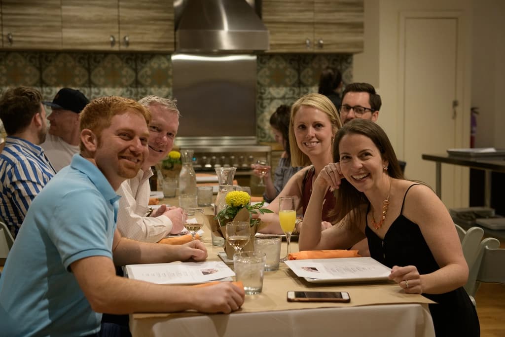 A group of people dining together at a table, smiling and enjoying their time in a warmly lit kitchen setting.