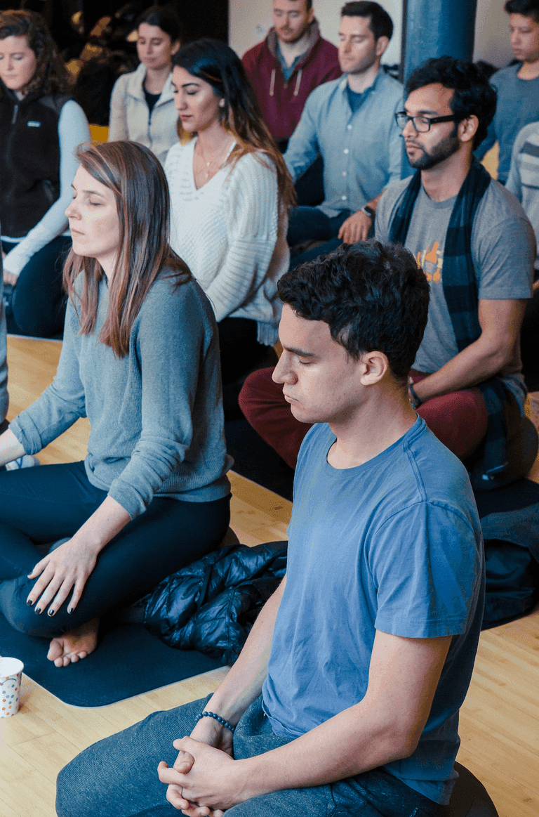People sitting cross-legged in a group meditation session, eyes closed and focused, on a wooden floor in a calm indoor space.