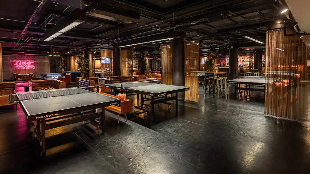 Spacious indoor venue featuring multiple ping pong tables, leather seating, and a neon sign, creating a relaxed and playful atmosphere.