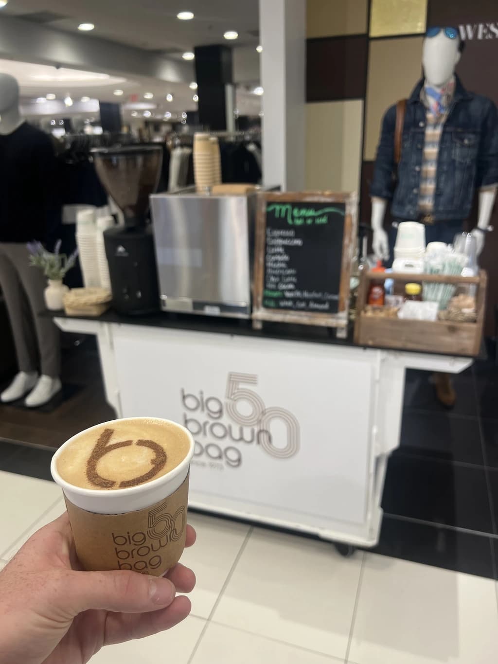 Espresso cart setup with a hand holding a latte cup featuring branded latte art. Background includes a “Big Brown Bag” sign.